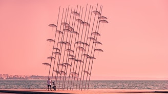 A sculpture with numerous tall metal poles holding umbrellas tilted at an angle by the seaside. The sky and water have a pink hue, and a person stands near the sculpture.