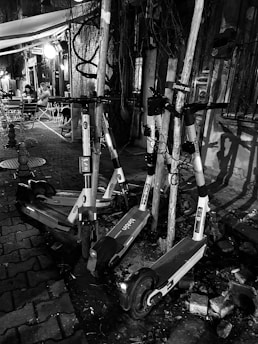 A group of electric scooters is parked haphazardly against poles on a cobblestone street. The setting appears urban, with a small outdoor seating area visible in the background. Two people are seated at a table under a canopy, and there is dim, ambient lighting from a nearby light fixture.