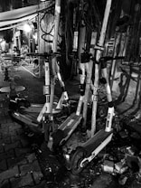 A group of electric scooters is parked haphazardly against poles on a cobblestone street. The setting appears urban, with a small outdoor seating area visible in the background. Two people are seated at a table under a canopy, and there is dim, ambient lighting from a nearby light fixture.