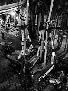 A group of electric scooters is parked haphazardly against poles on a cobblestone street. The setting appears urban, with a small outdoor seating area visible in the background. Two people are seated at a table under a canopy, and there is dim, ambient lighting from a nearby light fixture.