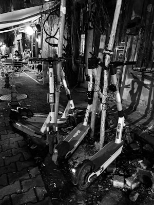 A group of electric scooters is parked haphazardly against poles on a cobblestone street. The setting appears urban, with a small outdoor seating area visible in the background. Two people are seated at a table under a canopy, and there is dim, ambient lighting from a nearby light fixture.