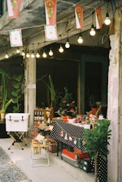 A small market stall is set up under strings of hanging lights. The table is covered with a black polka-dot cloth and decorated with Christmas-themed items such as miniature Santa hats. Various goods, including bottles, mugs, and small gifts, are displayed. A cooler sits on a stand nearby, while plants and old walls create a rustic atmosphere.