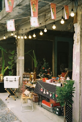 A small market stall is set up under strings of hanging lights. The table is covered with a black polka-dot cloth and decorated with Christmas-themed items such as miniature Santa hats. Various goods, including bottles, mugs, and small gifts, are displayed. A cooler sits on a stand nearby, while plants and old walls create a rustic atmosphere.