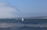 A tranquil seascape with a sailboat and a motorboat gliding across the water, accompanied by two kayaks in the foreground. The sky is clear with a slight haze in the distance, and the horizon features a low-lying coastal landscape.