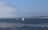 A tranquil seascape with a sailboat and a motorboat gliding across the water, accompanied by two kayaks in the foreground. The sky is clear with a slight haze in the distance, and the horizon features a low-lying coastal landscape.