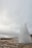 A geyser erupts in a natural landscape, sending a tall plume of water and steam into the air. The sky is overcast, and the ground appears rocky and wet. Vegetation can be seen in the background, along with distant hills.