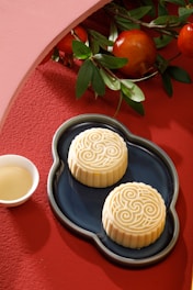 Two intricately designed mooncakes rest on a dark blue platter with a silver outline. A small white cup of tea accompanies them on a textured red surface. Above, there are branches with green leaves and vibrant red pomegranates, creating a harmonious blend of colors and textures.