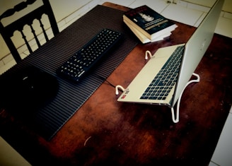 A laptop set on a table, elevated with a stand, alongside books and a keyboard. The table surface is wooden, and there is a black chair with a slatted back. The books are stacked on the right side, and the keyboard is placed on a textured mat.