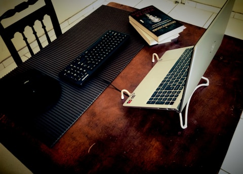 A laptop set on a table, elevated with a stand, alongside books and a keyboard. The table surface is wooden, and there is a black chair with a slatted back. The books are stacked on the right side, and the keyboard is placed on a textured mat.