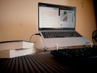 A laptop with an open webpage is on a desk, alongside two stacked books and a keyboard. The background is a plain wall, and a small fan is partially visible. The lighting creates a warm, cozy atmosphere.