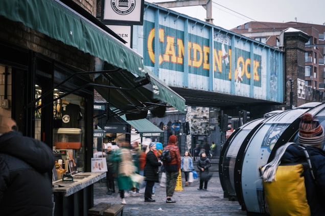 A bustling urban scene featuring a market area with people walking under a blue railway bridge. The bridge has large yellow letters reading 'Camden Lock'. On the left, there is a food stall with green awnings. The scene is lively with several people in winter clothing, suggesting a cold day.