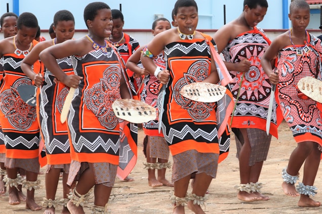 A group of women in traditional clothing are performing a coordinated dance. They are wearing colorful garments with intricate patterns, predominantly in orange and black. Each is holding a traditional shield and adorned with beaded jewelry. The ground is bare, and the setting appears to be outdoors in a bright environment.