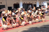 A group of performers engaged in a traditional dance is dressed in vibrant cultural attire. The performers wear fur embellishments and carry long sticks, executing synchronized high kicks. The background features a building with several observers watching the event.
