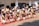 A group of performers engaged in a traditional dance is dressed in vibrant cultural attire. The performers wear fur embellishments and carry long sticks, executing synchronized high kicks. The background features a building with several observers watching the event.