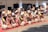 A group of performers engaged in a traditional dance is dressed in vibrant cultural attire. The performers wear fur embellishments and carry long sticks, executing synchronized high kicks. The background features a building with several observers watching the event.