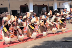 A group of performers engaged in a traditional dance is dressed in vibrant cultural attire. The performers wear fur embellishments and carry long sticks, executing synchronized high kicks. The background features a building with several observers watching the event.