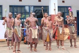 A group of people dressed in traditional attire featuring patterned skirts, beaded accessories, and holding sticks stand together in front of a building with large windows. They appear to be participating in a cultural or traditional event.