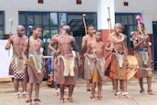 A group of people dressed in traditional attire featuring patterned skirts, beaded accessories, and holding sticks stand together in front of a building with large windows. They appear to be participating in a cultural or traditional event.