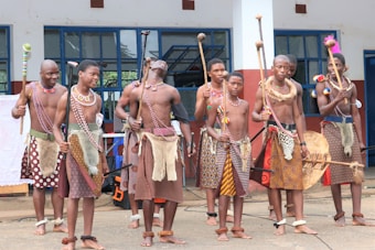 A group of people dressed in traditional attire featuring patterned skirts, beaded accessories, and holding sticks stand together in front of a building with large windows. They appear to be participating in a cultural or traditional event.