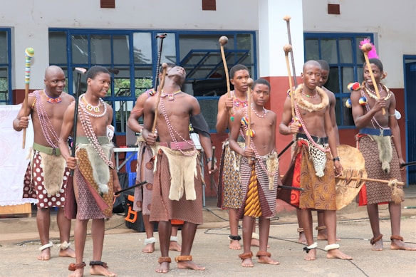 A group of people dressed in traditional attire featuring patterned skirts, beaded accessories, and holding sticks stand together in front of a building with large windows. They appear to be participating in a cultural or traditional event.