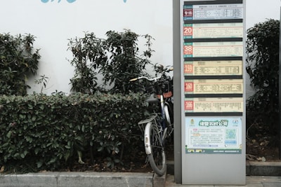A public transportation schedule is displayed on a tall metal stand next to a partially visible bicycle. The stand is surrounded by green shrubbery, giving an urban yet natural setting. The schedule contains multiple sections with text and numbers, likely indicating bus routes or times.