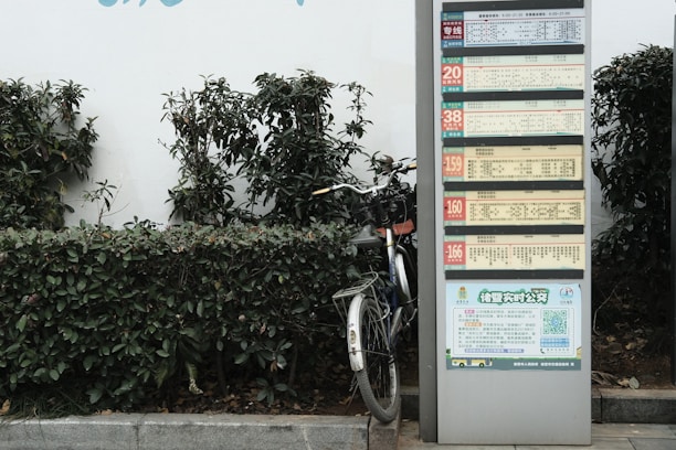 A public transportation schedule is displayed on a tall metal stand next to a partially visible bicycle. The stand is surrounded by green shrubbery, giving an urban yet natural setting. The schedule contains multiple sections with text and numbers, likely indicating bus routes or times.