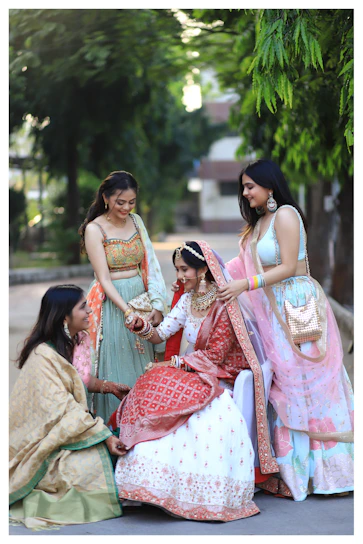 A group of young women laughing together, dressed in a mix of traditional sarees and lehengas with modern twists, set in a festive outdoor setting.