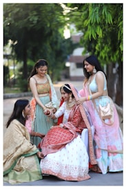 Four women are dressed in colorful traditional attire, likely for a celebration or wedding. The central figure is sitting, wearing a red and white lehenga with intricate designs and adorned with jewelry. The other three women are standing around her, wearing lehengas in pastel shades and also accessorized with jewelry. They appear joyful and engaged in a candid, cheerful moment outdoors with greenery and a building in the background.