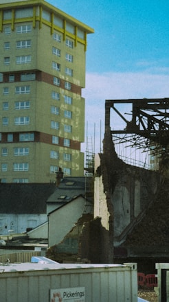 A partially demolished building stands next to a tall residential tower. The tower has yellow accents and multiple windows, while the demolished structure is surrounded by rubble and construction materials. There are construction signs and temporary fences around the area with clear skies above.