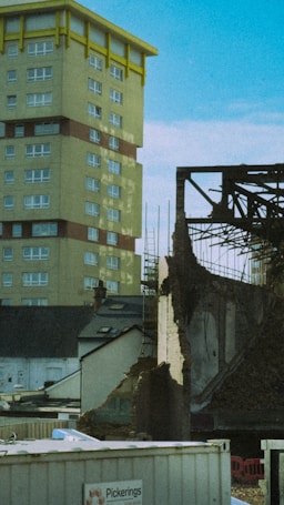 A partially demolished building stands next to a tall residential tower. The tower has yellow accents and multiple windows, while the demolished structure is surrounded by rubble and construction materials. There are construction signs and temporary fences around the area with clear skies above.
