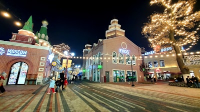 A festive urban street scene features illuminated buildings and a large, decorated tree with string lights. The street is populated with people enjoying the atmosphere, including children wearing winter clothing. Various shops and restaurants are visible, such as an ice cream shop labeled 'Moscow' and a building with the sign 'EVOS'. The setting suggests a busy, lively environment.