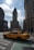 A classic yellow taxi is driving through a New York City intersection with the iconic Flatiron Building prominently in the background. The street is lined with tall, historic buildings under a partly cloudy sky, with sunlight casting shadows on the street.