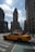 A classic yellow taxi is driving through a New York City intersection with the iconic Flatiron Building prominently in the background. The street is lined with tall, historic buildings under a partly cloudy sky, with sunlight casting shadows on the street.