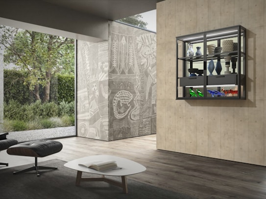 A modern interior space featuring a minimalist lounge area with a black leather chair and footrest beside a white, low-profile coffee table. The room has large glass walls offering a view of lush greenery outside. On the right, there is a decorative wall with intricate mural-like patterns, and a sleek, black-framed display cabinet on it. The cabinet contains various artifacts and ceramic vases, both with neutral and vibrant colors.