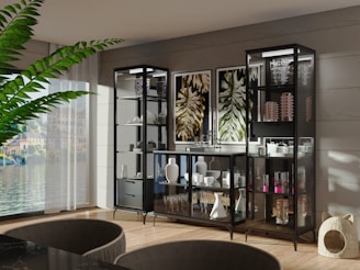 A modern, elegant living room features a large plant, a wall with multiple glass cabinets displaying decorative items like vases and glasses, and artwork depicting abstract leaves. Light streams in through a large window, illuminating the wooden floor and sleek furniture.