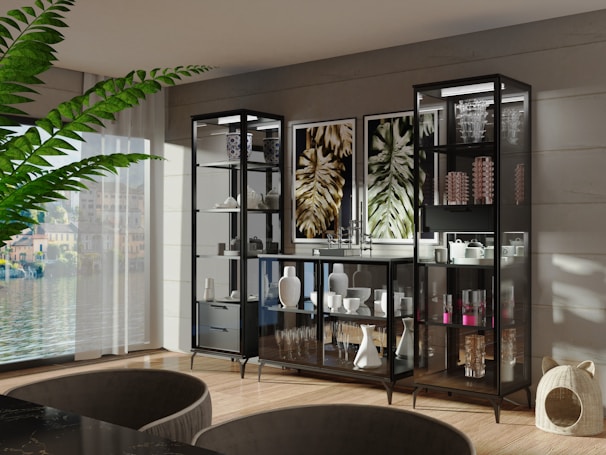 A modern, elegant living room features a large plant, a wall with multiple glass cabinets displaying decorative items like vases and glasses, and artwork depicting abstract leaves. Light streams in through a large window, illuminating the wooden floor and sleek furniture.