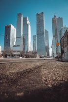 A view of tall, modern skyscrapers with reflective glass facades set against a clear blue sky. The photo is taken from a low angle on an urban street, with construction barriers and traffic signs visible. The street surface shows textured pavements and asphalt.