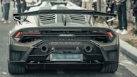 A high-performance sports car, characterized by its sleek design and aerodynamic features, is parked on a city street. The focus is on the rear of the car, showcasing dual exhaust pipes, a prominent spoiler, and distinctive tail lights. A crowd of people stands nearby, admiring the vehicle.