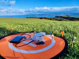 A small drone is placed on an orange landing pad with a smartphone nearby, situated on a green grassy field. In the background, there's a scenic view of the coastline with cliffs and a clear blue sky filled with clouds.