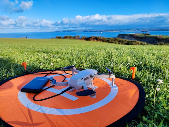 A small drone is placed on an orange landing pad with a smartphone nearby, situated on a green grassy field. In the background, there's a scenic view of the coastline with cliffs and a clear blue sky filled with clouds.