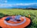 A small drone is placed on an orange landing pad with a smartphone nearby, situated on a green grassy field. In the background, there's a scenic view of the coastline with cliffs and a clear blue sky filled with clouds.