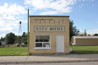 A small, rectangular brick building with a sign reading 'City Office' is situated next to a grassy area. The structure has a large window and a single door. To the left, a red-topped metal tower is visible, surrounded by trees and greenery. In the background, there are more structures and a backdrop of open skies with fluffy clouds.