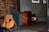 A wooden acoustic guitar is leaning against an amplifier, with a strap draped over it. Beside the guitar, there is an upright bass lying on its side on a wooden floor. The background features a textured wall with wooden paneling and framed pictures.