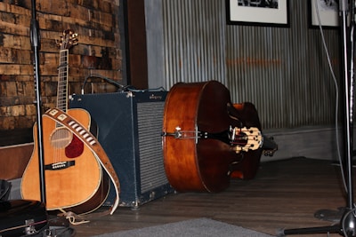 A wooden acoustic guitar is leaning against an amplifier, with a strap draped over it. Beside the guitar, there is an upright bass lying on its side on a wooden floor. The background features a textured wall with wooden paneling and framed pictures.