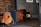 A wooden acoustic guitar is leaning against an amplifier, with a strap draped over it. Beside the guitar, there is an upright bass lying on its side on a wooden floor. The background features a textured wall with wooden paneling and framed pictures.