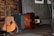 A wooden acoustic guitar is leaning against an amplifier, with a strap draped over it. Beside the guitar, there is an upright bass lying on its side on a wooden floor. The background features a textured wall with wooden paneling and framed pictures.