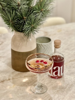 A marble table adorned with a cocktail glass filled with a pinkish liquid and red cherries. Next to it is a small bottle with a red label and a large vase containing green pine branches. The vase has a gradient design ranging from white to brown.