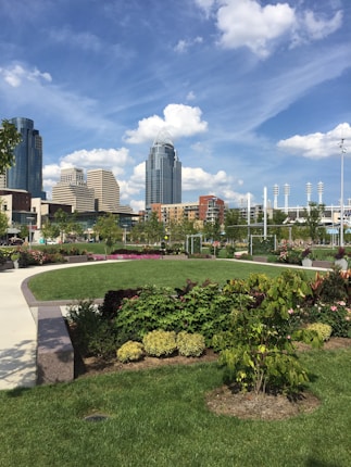 A vibrant urban park scene with lush green grass, colorful flower beds, and various plants. In the background, a skyline featuring modern skyscrapers, including one with a distinct architectural crown, and a cloudy blue sky above. The park features curved walkways and is surrounded by tall buildings.