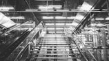 A modern indoor staircase with metal railings and steps, reflecting an industrial aesthetic. The surroundings include glass panels and bright, linear overhead lights that create a dynamic and futuristic atmosphere.