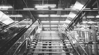A modern indoor staircase with metal railings and steps, reflecting an industrial aesthetic. The surroundings include glass panels and bright, linear overhead lights that create a dynamic and futuristic atmosphere.